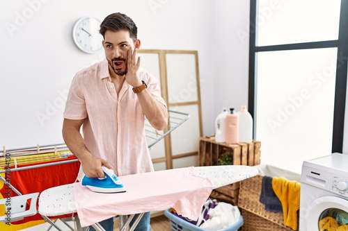 Young man with beard ironing clothes at home hand on mouth telling secret rumor  whispering malicious talk conversation