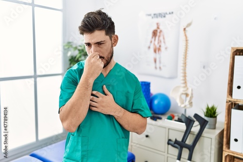 Young man with beard working at pain recovery clinic smelling something stinky and disgusting, intolerable smell, holding breath with fingers on nose. bad smell