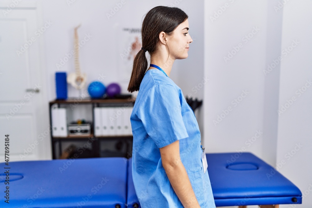 Young hispanic woman wearing physiotherapist uniform standing at clinic looking to side, relax profile pose with natural face and confident smile.