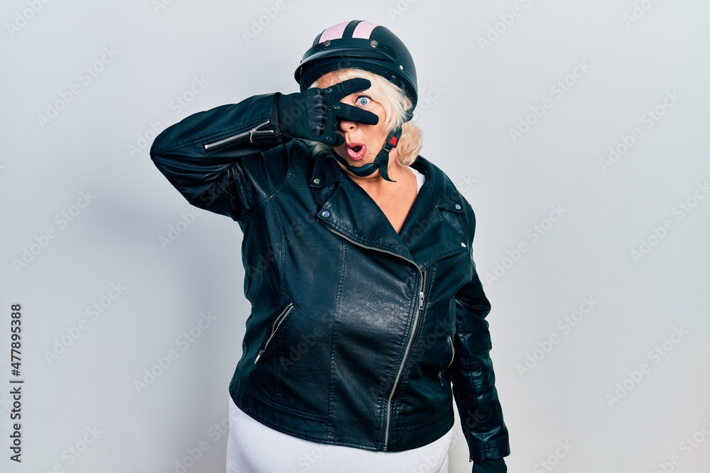 Middle age blonde woman holding motorcycle helmet peeking in shock covering face and eyes with hand, looking through fingers with embarrassed expression. Middle age blonde woman holding motorcycle helmet peeking in shock covering face and eyes with hand, looking through fingers with embarrassed expression.