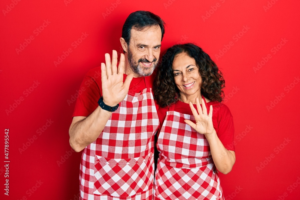 Middle age couple of hispanic woman and man wearing cook apron waiving saying hello happy and smiling, friendly welcome gesture Middle age couple of hispanic woman and man wearing cook apron waiving saying hello happy and smiling, friendly welcome gesture