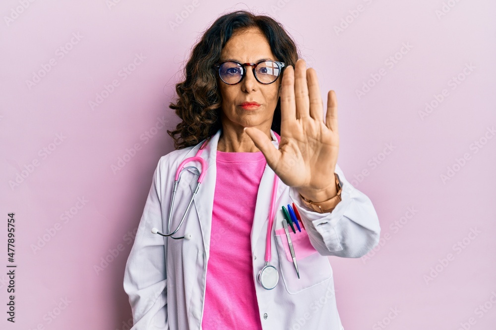 Middle age hispanic woman wearing doctor uniform and glasses doing stop sing with palm of the hand. warning expression with negative and serious gesture on the face.