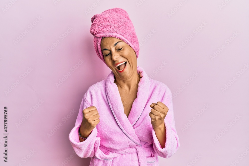 Middle age hispanic woman wearing shower towel cap and bathrobe very happy and excited doing winner gesture with arms raised, smiling and screaming for success. celebration concept.