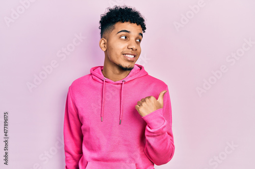 Young african american man wearing casual sweatshirt smiling with happy face looking and pointing to the side with thumb up.