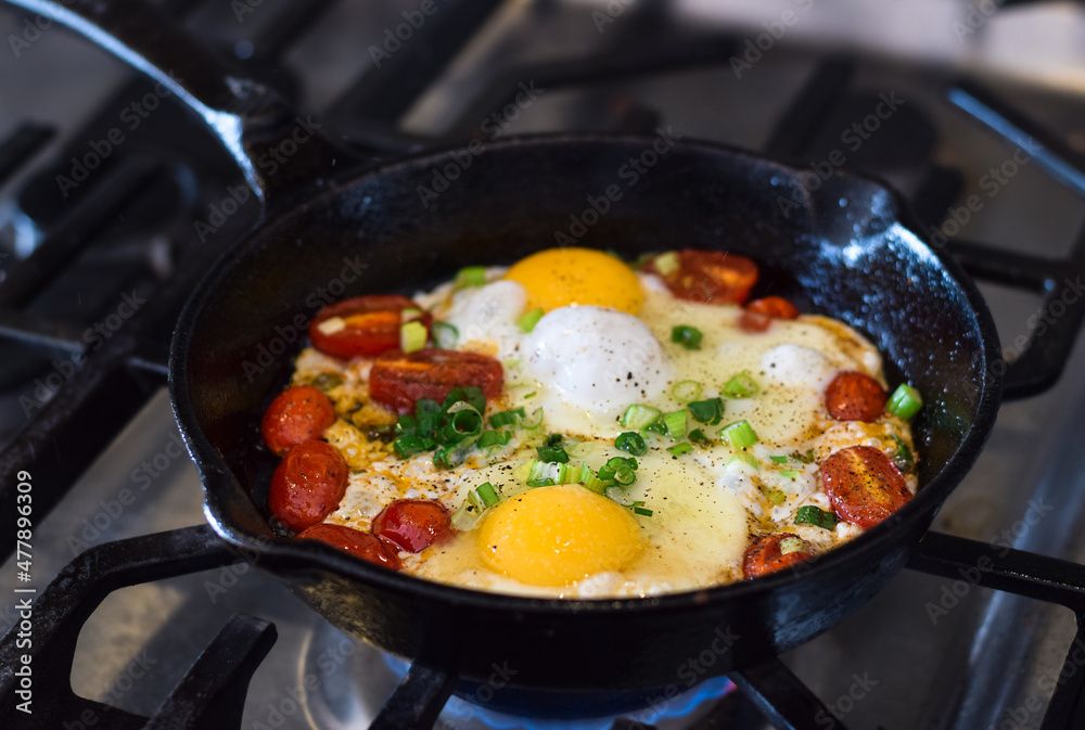 Breakfast eggs with tomatoes and green onions frying in cast iron pan on gas stove Breakfast eggs with tomatoes and green onions frying in cast iron pan on gas stove
