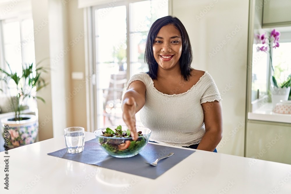 Young hispanic woman eating healthy salad at home smiling friendly offering handshake as greeting and welcoming. successful business.