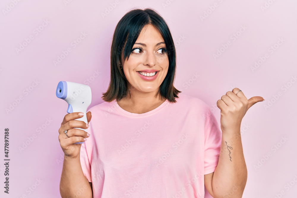 Young hispanic woman holding thermometer pointing thumb up to the side smiling happy with open mouth Young hispanic woman holding thermometer pointing thumb up to the side smiling happy with open mouth