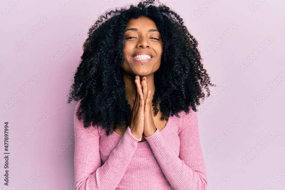 African american woman with afro hair wearing casual pink shirt praying with hands together asking for forgiveness smiling confident. African american woman with afro hair wearing casual pink shirt praying with hands together asking for forgiveness smiling confident.