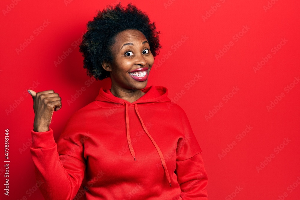 Young african american woman wearing casual sweatshirt smiling with happy face looking and pointing to the side with thumb up. Young african american woman wearing casual sweatshirt smiling with happy face looking and pointing to the side with thumb up.