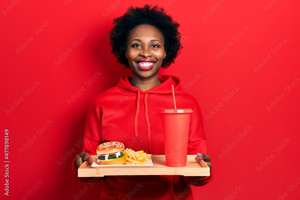 Young african american woman eating a tasty classic burger with fries and soda smiling with a happy and cool smile on face. showing teeth. Young african american woman eating a tasty classic burger with fries and soda smiling with a happy and cool smile on face. showing teeth.
