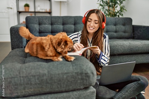 Young hispanic woman using laptop studying sitting on floor with dog at home