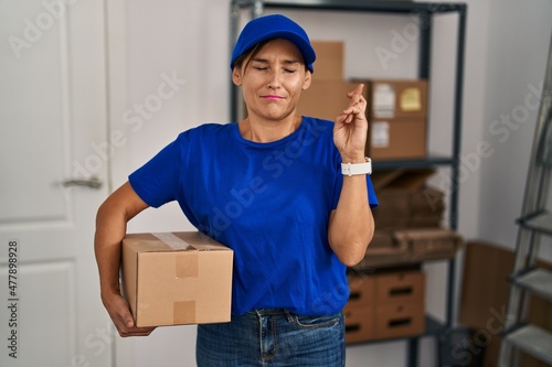 Middle age brunette woman working wearing delivery uniform and cap gesturing finger crossed smiling with hope and eyes closed. luck and superstitious concept.