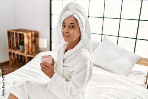 Middle age grey-haired woman drinking coffee sitting on bed at bedroom