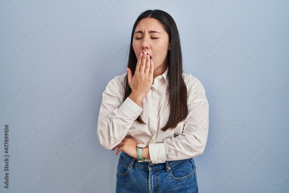 Young latin woman standing over blue background bored yawning tired covering mouth with hand. restless and sleepiness. Young latin woman standing over blue background bored yawning tired covering mouth with hand. restless and sleepiness.