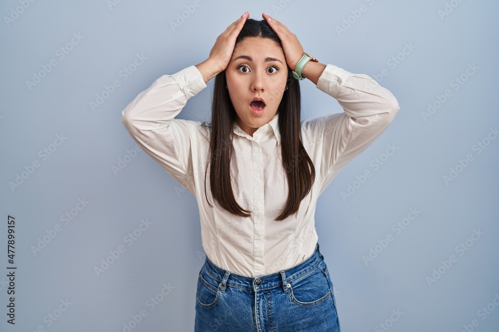 Young latin woman standing over blue background crazy and scared with hands on head, afraid and surprised of shock with open mouth