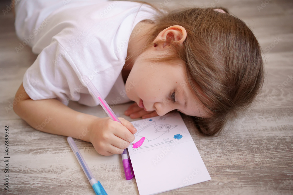 Homeschooled girl, draws in a notebook lying on the floor, dressed in a white T-shirt. High quality photo Homeschooled girl, draws in a notebook lying on the floor, dressed in a white T-shirt. High quality photo
