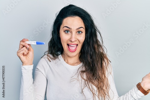 Young hispanic woman holding thermometer celebrating achievement with happy smile and winner expression with raised hand