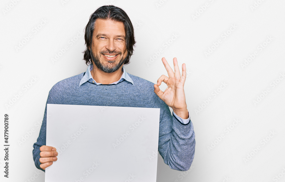 Middle age handsome man holding blank empty banner doing ok sign with fingers, smiling friendly gesturing excellent symbol