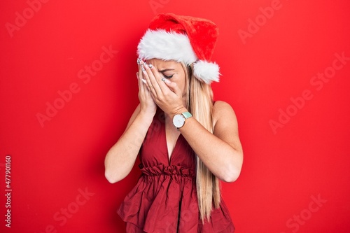 Young blonde girl wearing christmas hat with sad expression covering face with hands while crying. depression concept.
