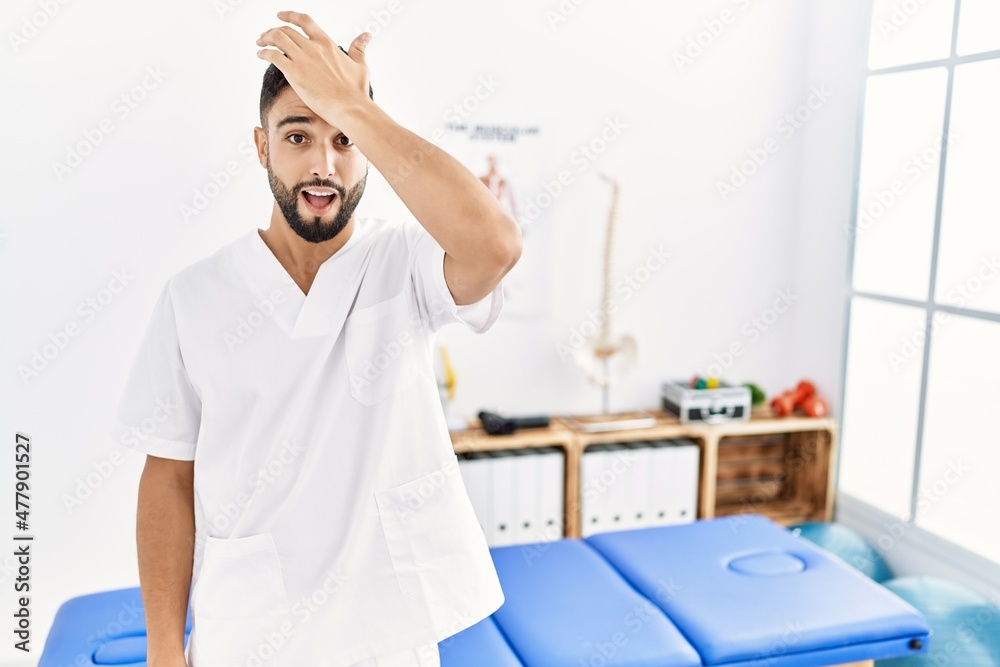Young handsome man with beard working at pain recovery clinic surprised with hand on head for mistake, remember error. forgot, bad memory concept. Young handsome man with beard working at pain recovery clinic surprised with hand on head for mistake, remember error. forgot, bad memory concept.
