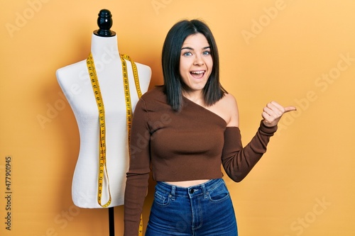 Young brunette woman standing by manikin pointing thumb up to the side smiling happy with open mouth