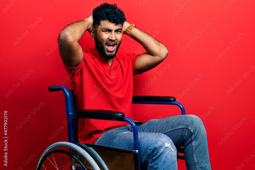Arab man with beard sitting on wheelchair crazy and scared with hands on head, afraid and surprised of shock with open mouth Arab man with beard sitting on wheelchair crazy and scared with hands on head, afraid and surprised of shock with open mouth