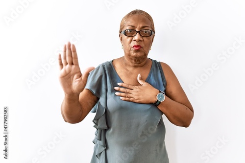 Mature hispanic woman wearing glasses standing over isolated background swearing with hand on chest and open palm, making a loyalty promise oath
