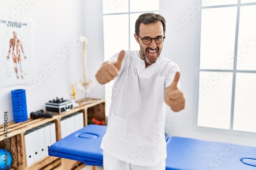 Middle age man with beard working at pain recovery clinic approving doing positive gesture with hand  thumbs up smiling and happy for success. winner gesture.
