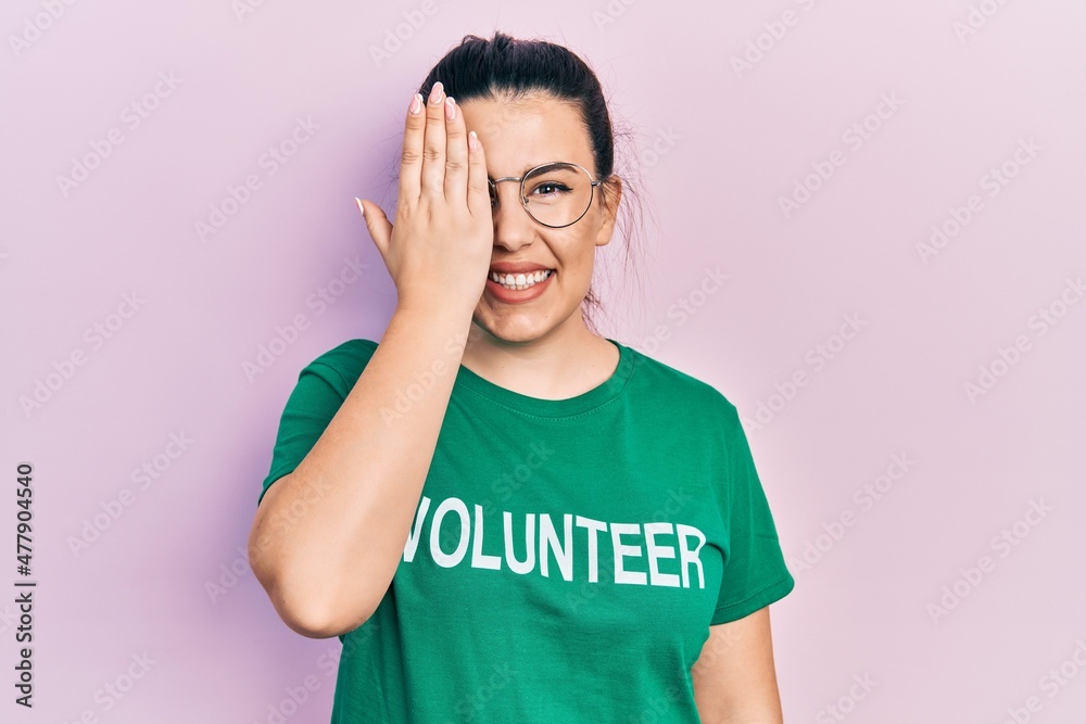 Young hispanic woman wearing volunteer t shirt covering one eye with hand, confident smile on face and surprise emotion. Young hispanic woman wearing volunteer t shirt covering one eye with hand, confident smile on face and surprise emotion.