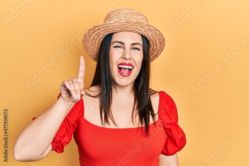 Young hispanic woman wearing summer hat showing and pointing up with finger number one while smiling confident and happy.