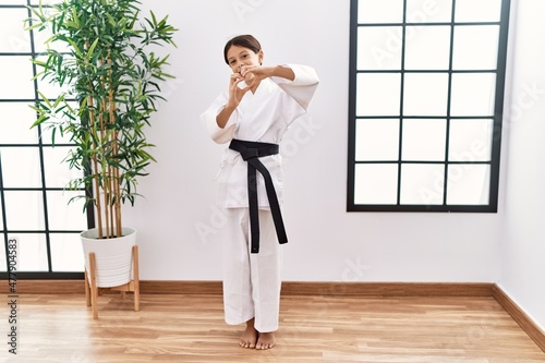 Young hispanic girl wearing karate kimono and black belt smiling in love doing heart symbol shape with hands. romantic concept.