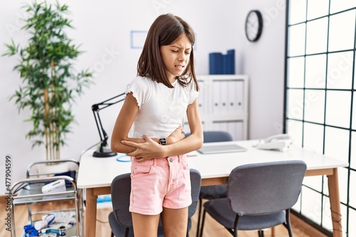 Foto Young hispanic girl standing at pediatrician clinic with hand on stomach because indigestion, painful illness feeling unwell