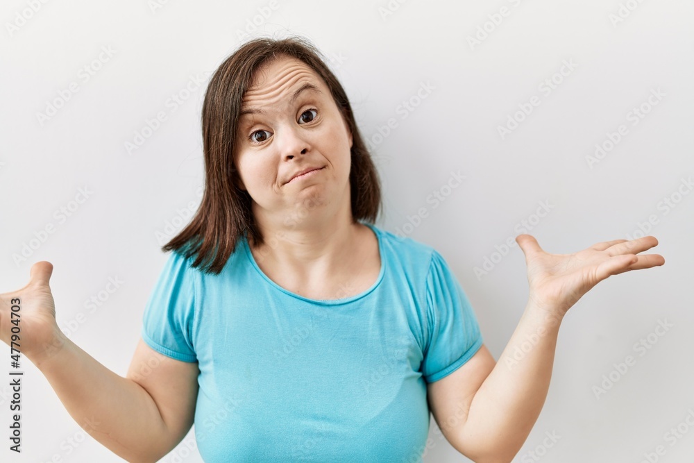 Young down syndrome woman standing over isolated background clueless and confused expression with arms and hands raised. doubt concept. Young down syndrome woman standing over isolated background clueless and confused expression with arms and hands raised. doubt concept.