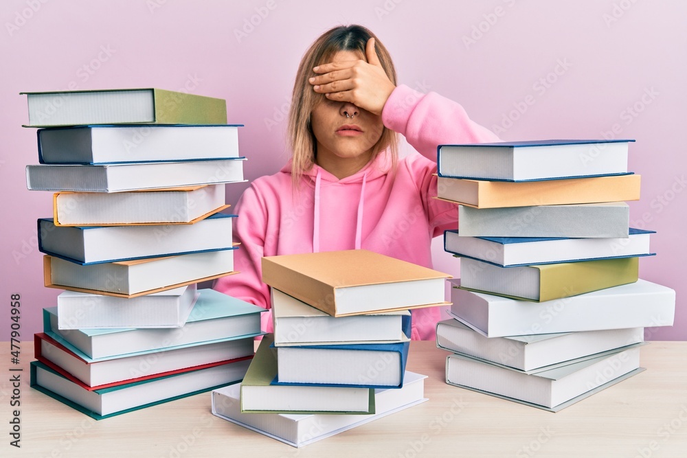 Young caucasian woman sitting on the table with books covering eyes with hand, looking serious and sad. sightless, hiding and rejection concept Young caucasian woman sitting on the table with books covering eyes with hand, looking serious and sad. sightless, hiding and rejection concept