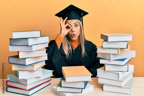 Young caucasian woman wearing graduation ceremony robe sitting on the table doing ok gesture shocked with surprised face, eye looking through fingers. unbelieving expression.