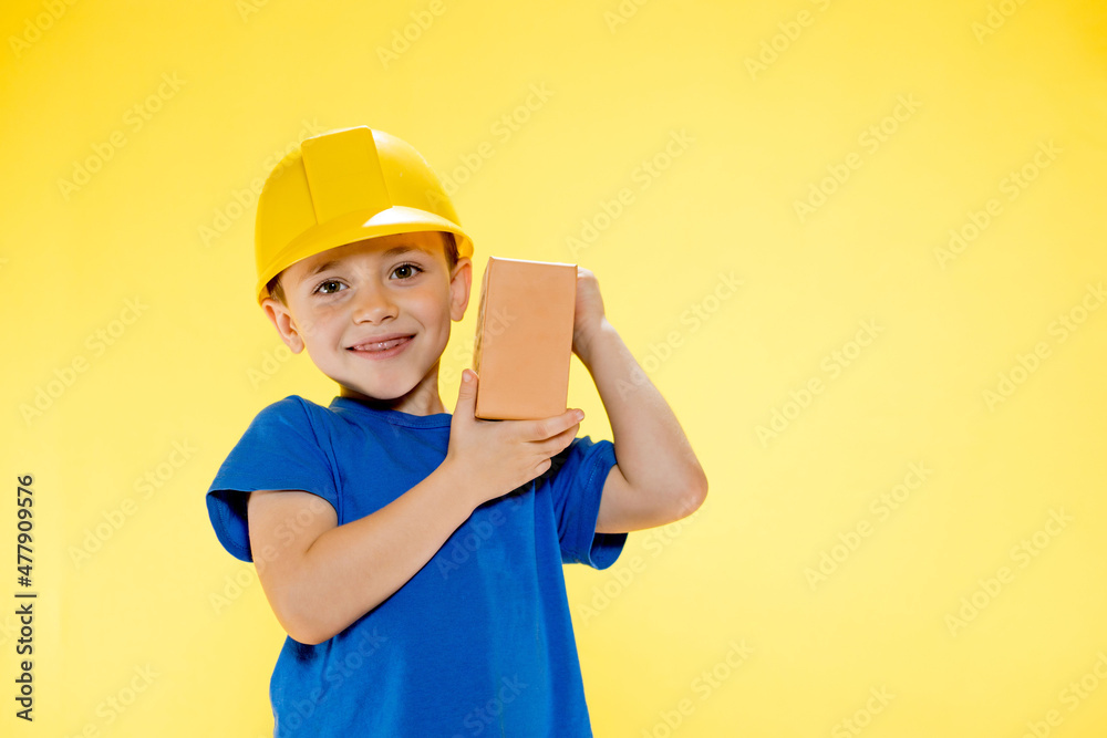 Boy in a construction helmet holds a brick in his hands on yellow background Boy in a construction helmet holds a brick in his hands on yellow background