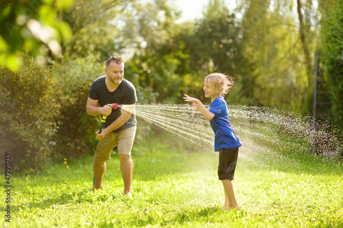 Funny little boy with his father playing with garden hose in sunny backyard. Preschooler child having fun with spray of water. Summer outdoors activity for kids.