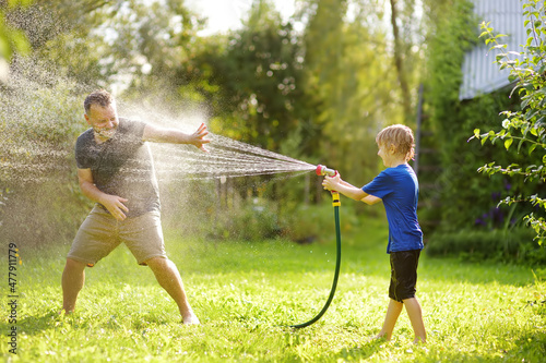 Funny little boy with his father playing with garden hose in sunny backyard. Preschooler child having fun with spray of water. Summer outdoors activity for kids. photo