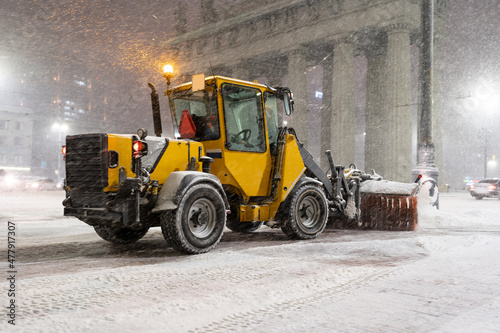 Winter service vehicle clearing thoroughfares of ice and snow, yellow snow plow tractor working on empty street at night in heavy snowfall to keep roads clear and safe for driving during winter