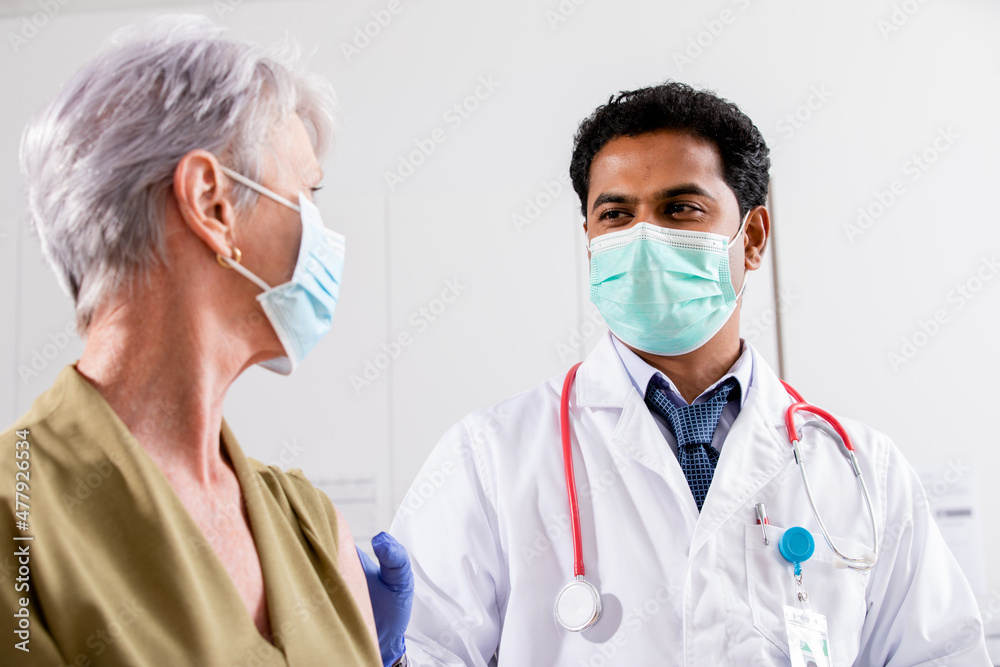 A Young Indian Male Medical Doctor Consults with an Elderly Senior Female Patient about Covid-19 Vaccine Injection Wearing Generic ID Badge, Lab Coat, Gloves and Mask in Hospital or Health Clinic. A Young Indian Male Medical Doctor Consults with an Elderly Senior Female Patient about Covid-19 Vaccine Injection Wearing Generic ID Badge, Lab Coat, Gloves and Mask in Hospital or Health Clinic.