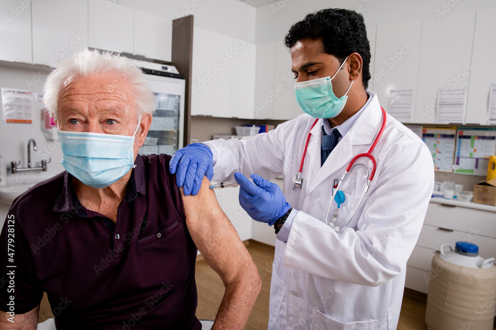 A Young Indian Male Medical Doctor Administering a Covid-19 Vaccine with a Syringe Needle to an Elderly Senior Male Patient Wearing Generic ID Badge, Gloves and Mask in Hospital or Health Clinic. A Young Indian Male Medical Doctor Administering a Covid-19 Vaccine with a Syringe Needle to an Elderly Senior Male Patient Wearing Generic ID Badge, Gloves and Mask in Hospital or Health Clinic.