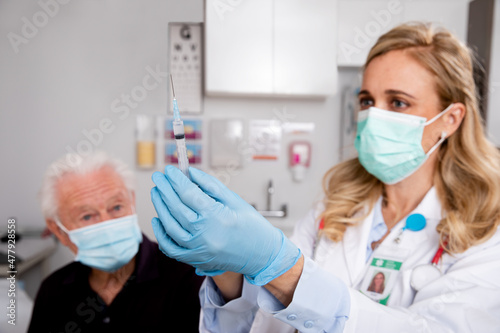 A Young White Female Medical Doctor Administering a Covid-19 Vaccine with a Syringe Needle to an Elderly Senior Male Patient Wearing Generic ID Badge  Gloves and Mask in Hospital or Health Clinic.