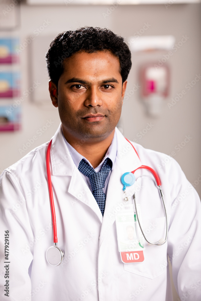 A Portrait of an Young Indian Male Medical Doctor with Generic ID Badge wearing a Lab Coat and Stethoscope in a Hospital or Health Clinic. Mid Shot. A Portrait of an Young Indian Male Medical Doctor with Generic ID Badge wearing a Lab Coat and Stethoscope in a Hospital or Health Clinic. Mid Shot.