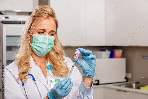A Young Female Medical Doctor Extracting a Covid-19 Vaccine with a Syringe Needle Wearing Generic ID Badge, Gloves, Mask, White Lab Coat and Stethoscope in Hospital or Health Clinic.