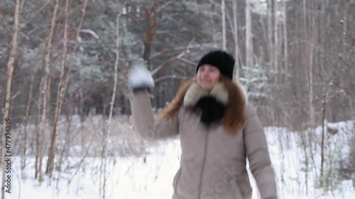 Red-haired girl throws snowballs, smiles and laughs in winter forest in warm clothes: black hat and light jacket, 4k. Blurred forest background
