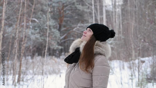 Portrait of a red-haired girl in warm clothes: black hat and light jacket in winter forest looks around, 4k. Blurred forest background
