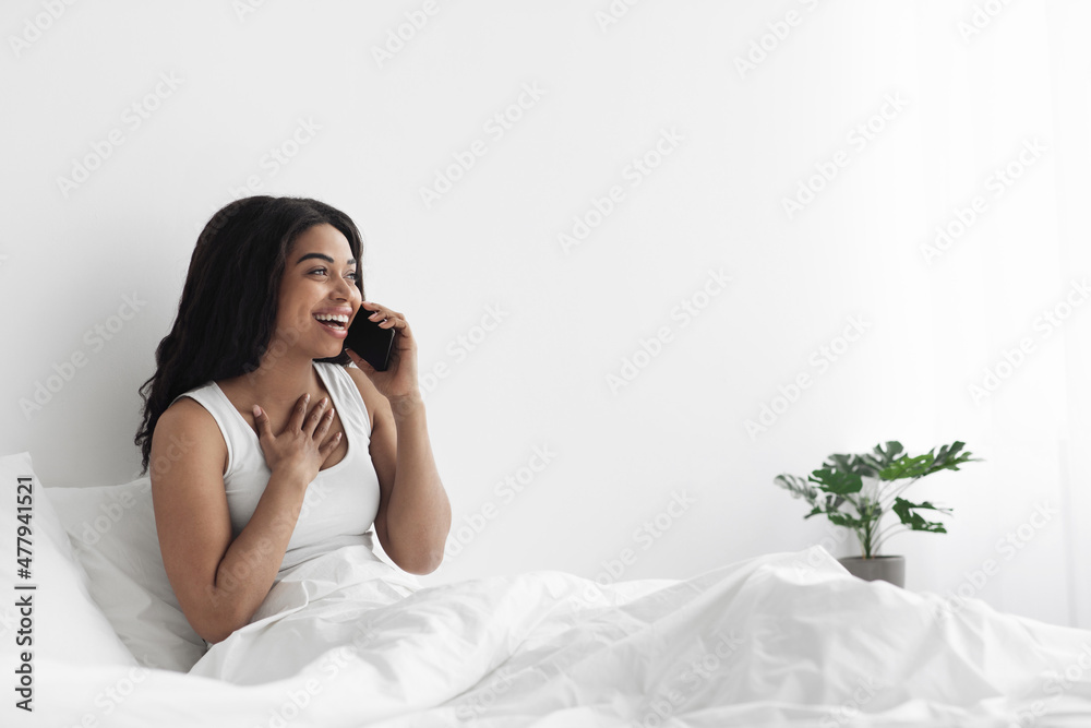 Happy african american lady sitting on bed and talking on smartphone, looking at copy space, resting in morning at home Happy african american lady sitting on bed and talking on smartphone, looking at copy space, resting in morning at home