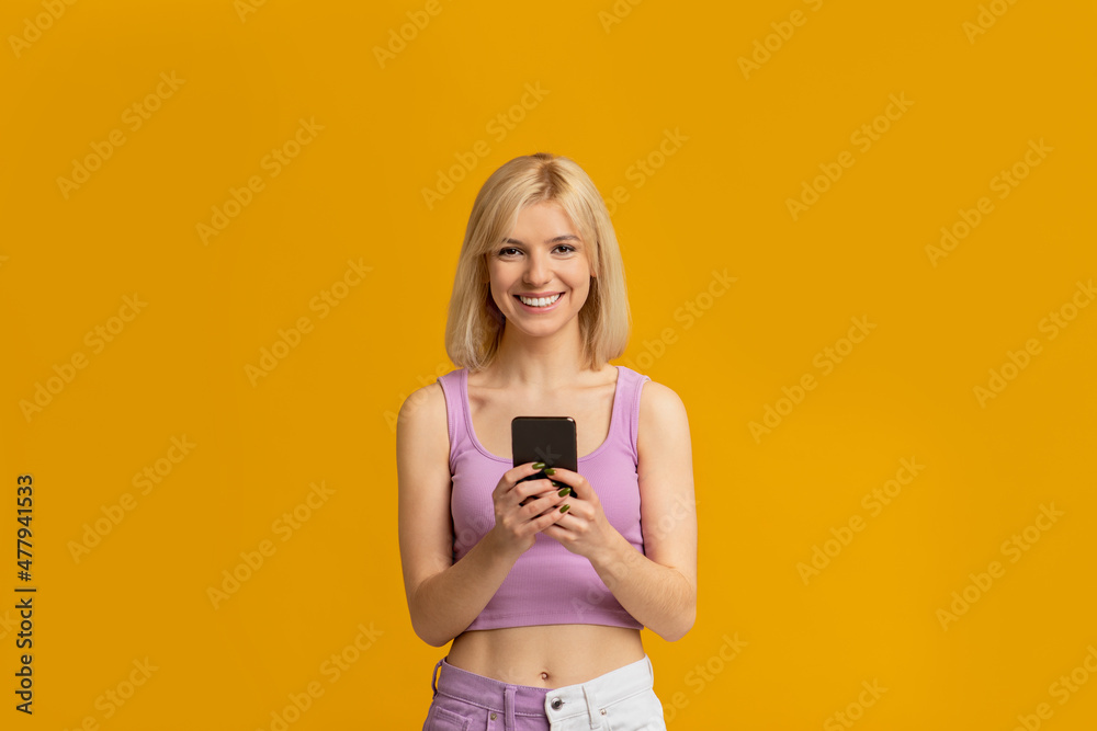 Great app. Positive young woman holding and using cellphone, chatting online isolated on yellow studio background Great app. Positive young woman holding and using cellphone, chatting online isolated on yellow studio background
