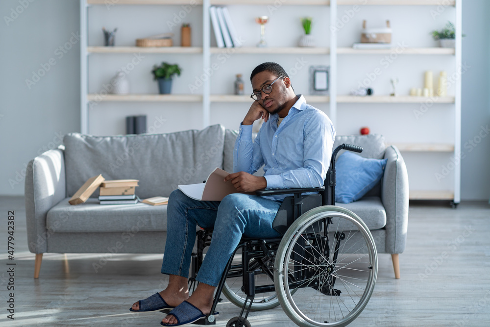 Impaired black man in wheelchair reading notebook, studying or working from home, full length Impaired black man in wheelchair reading notebook, studying or working from home, full length