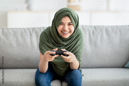 Portrait of happy teen girl in hijab holding joystick, playing videogame at home. Lockdown pastimes and hobbies